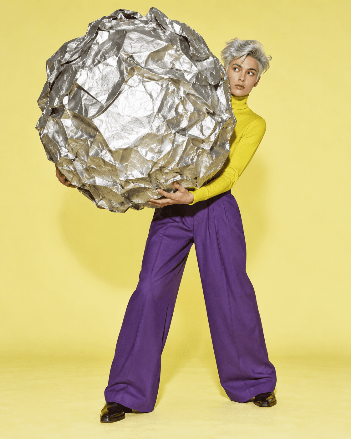 Surreal editorial artwork: a person in yellow peers around a giant crumpled silver paper sphere.