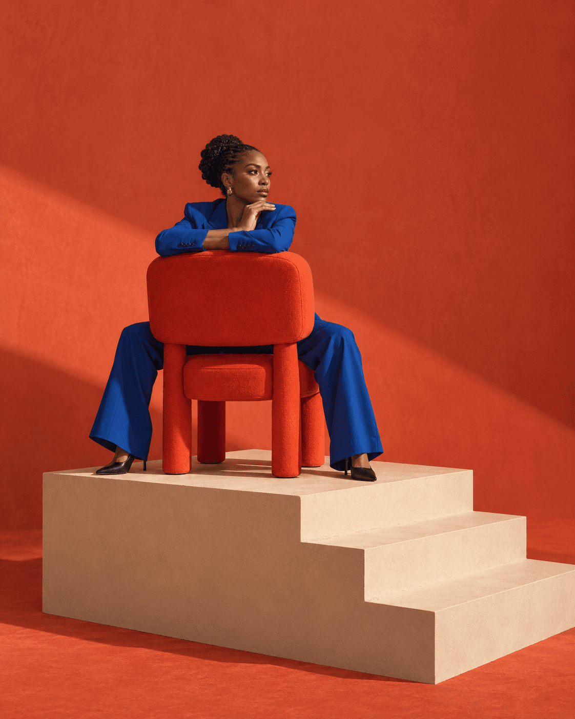 Surreal editorial artwork: a thoughtful person in blue sits backwards on an oversized red chair on a stepped platform.