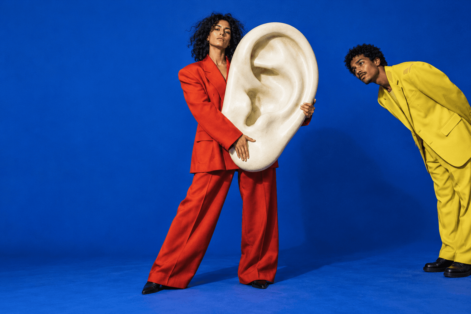 Surreal editorial artwork: a person in red holds an enormous sculptural ear while another person leans into the frame.